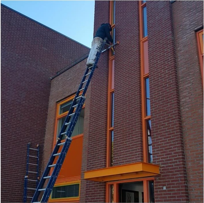 Schilder op een ladder bij oranje kozijnen van een bakstenen pand