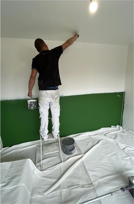 Schilder van GMH op een trap die een plafond wit schildert, de wand eronder donkergroen