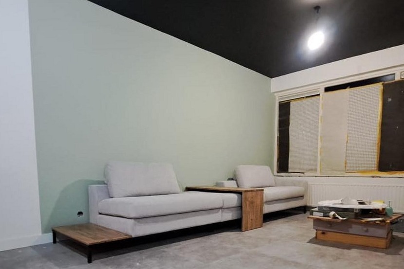 Woonkamer met een pas geschilderde zachtgroene wand, een zwart plafond en afgeplakte ramen, met een lichte bank tegen de wand